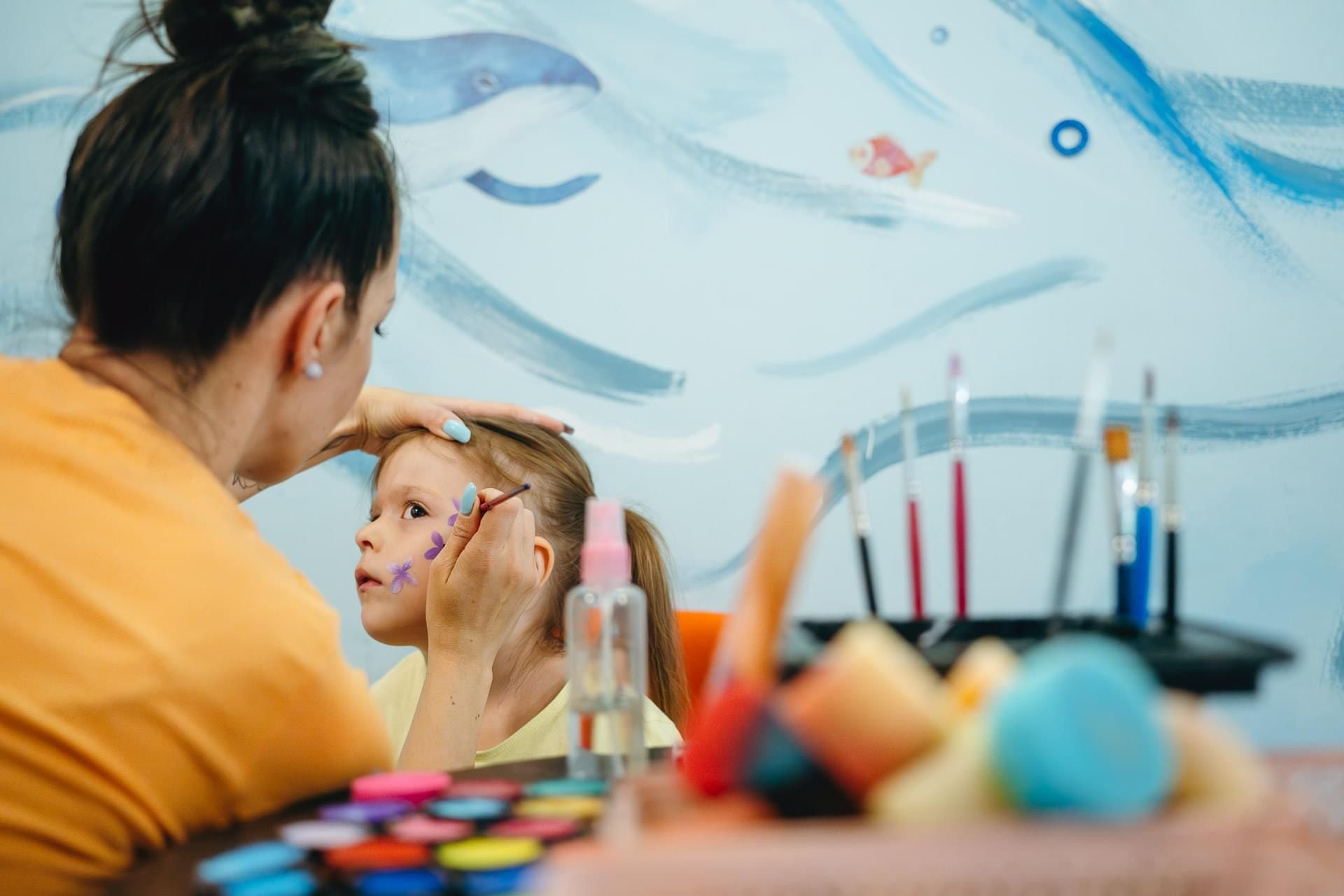 Woman painting child's face with purple flowers, brushes and paints nearby, aquatic mural in background.