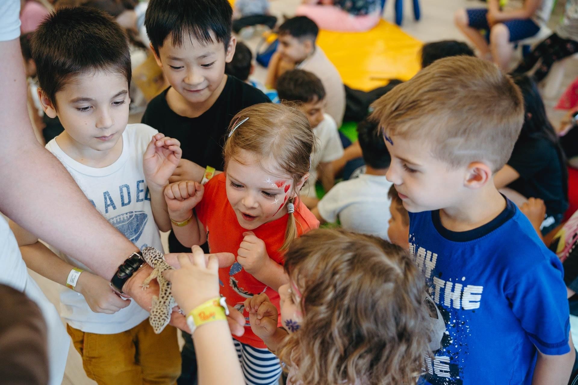 Group of children excitedly observing a gecko in an adult's hand during an indoor activity.
