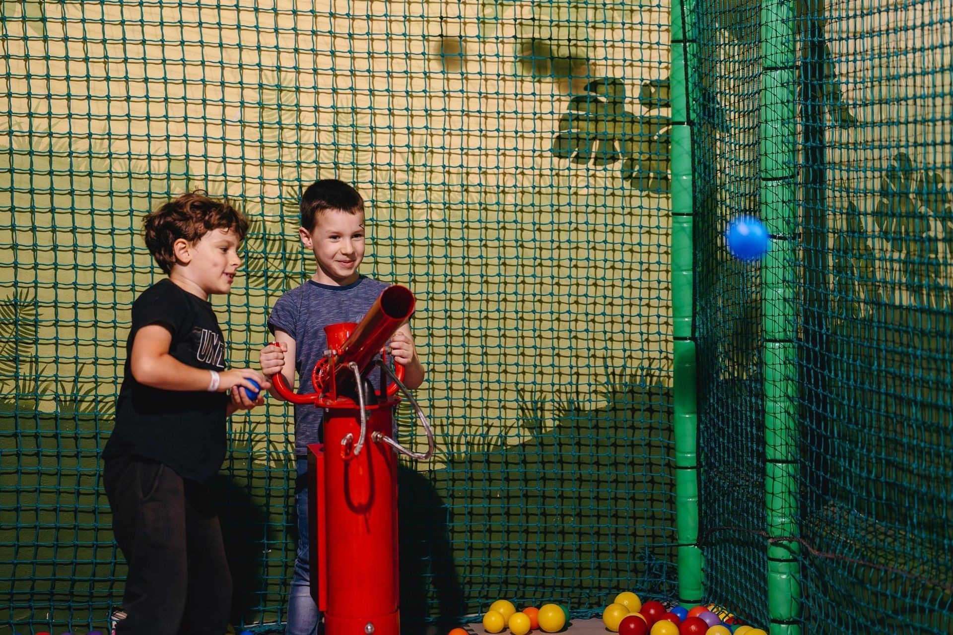 Two children playing with a red ball cannon in a netted play area filled with colorful balls.