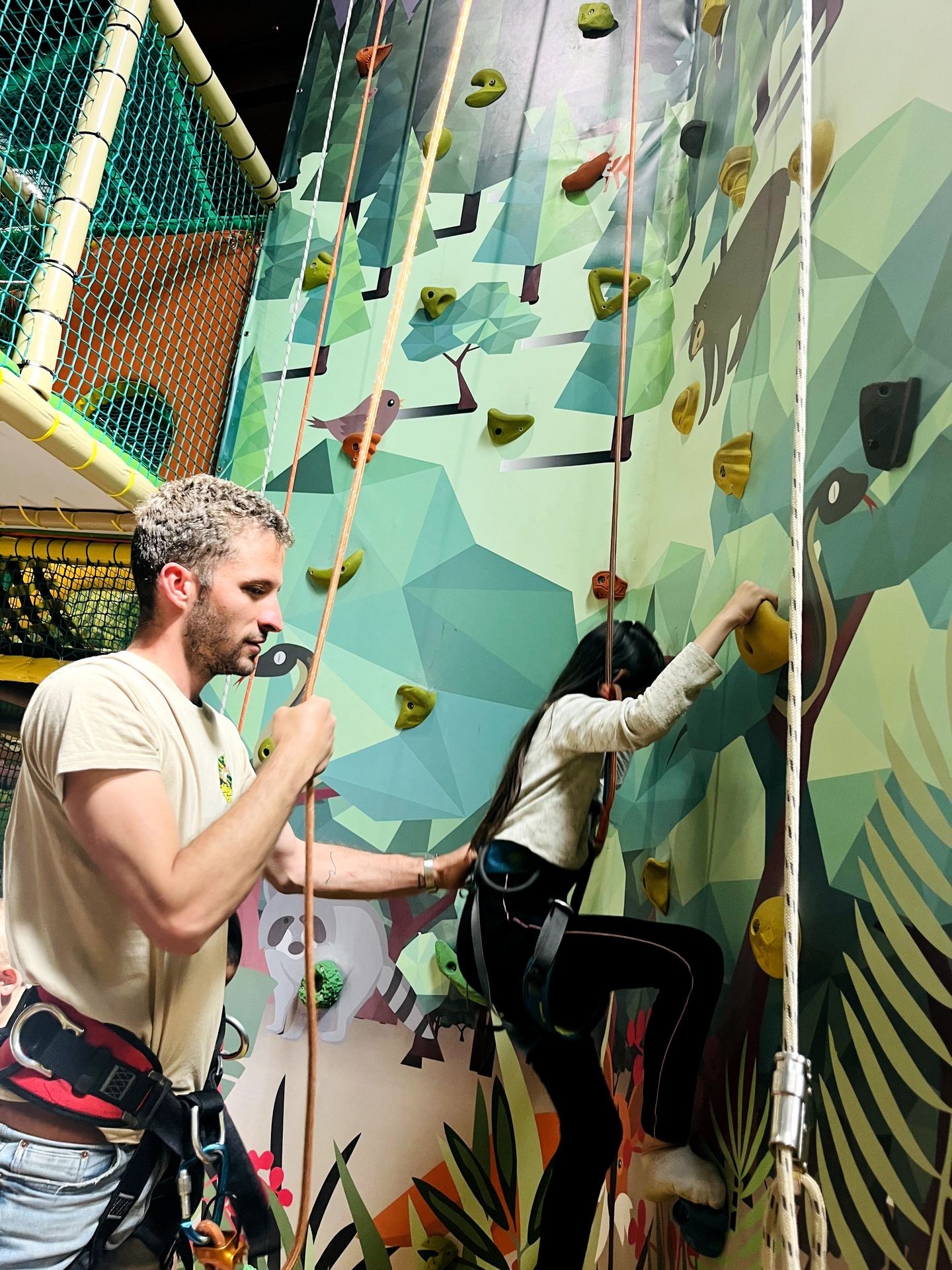 Man assisting a woman climbing an indoor rock wall with a colorful nature-themed design.