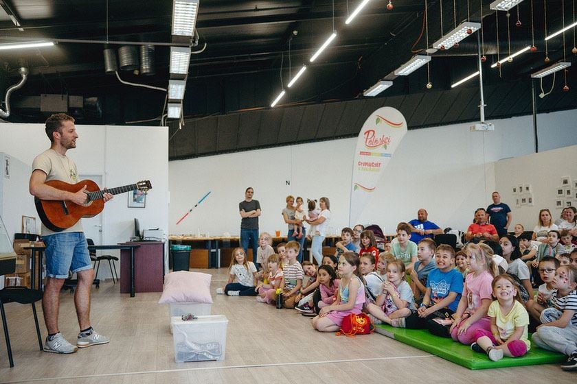 Man playing guitar in front of a group of children seated on the floor in a classroom setting.